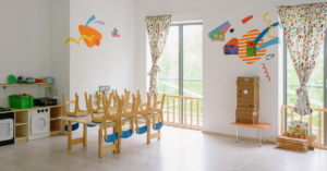 Bright Montessori-style childcare classroom with wooden tables and chairs, play kitchen, learning materials and natural light from large windows.
