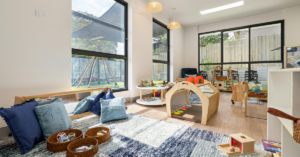Modern Montessori childcare classroom with natural wooden play equipment, soft floor seating and large windows overlooking outdoor play area.