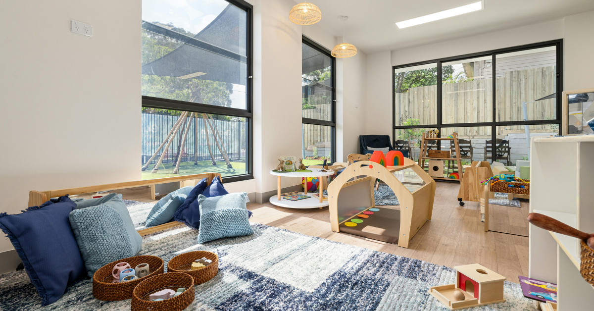 Modern Montessori childcare classroom with natural wooden play equipment, soft floor seating and large windows overlooking outdoor play area.